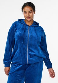 Blauer Velours-Trainingsanzug mit einer Kapuzenjacke mit Reißverschluss und passenden Hosen, geprägtem Sternenmuster, elastischen Bündchen und entspannter Passform.