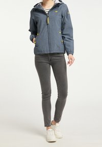 Blå lättviktig jacka med huva, med gula knappar och dragkedja, bärs över grå jeans och vita sneakers.