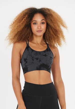 Kvinde med krøllet hår iført en sort tie-dye sports-bh og sorte højtaljede leggings, stående mod en ensfarvet hvid baggrund.