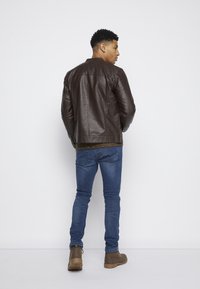 Chaqueta de cuero marrón con una textura suave, puños acanalados y cierre de cremallera, combinada con jeans azules y botas marrones. Modelo de espaldas.