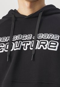 Černá mikina s kapucí a stahovací šňůrkou, na přední straně výrazný bílý nápis "VERSACE JEANS COUTURE". Měkká textura látky.