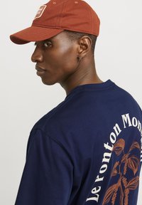 Marine t-skjorte med stort baktrykk av plante-design og teksten "Le Ponton Montaigne." Brun caps med brodert logo, avslappet passform.