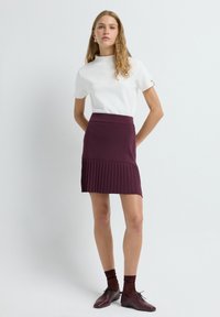 Weißes kurzärmliges Oberteil kombiniert mit einem burgunderroten plissierten Minirock. Der Rock verfügt über einen glatten Bund und vertikale Falten. Braune Schuhe und Socken runden den Look ab.