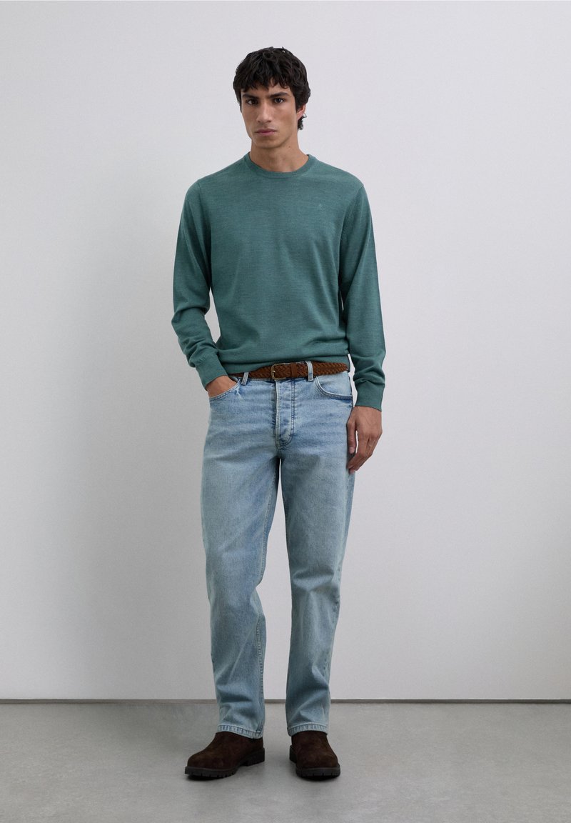 Teal långärmad tröja, ljusblå denimjeans och brunt texturerat bälte. Modellen står avslappnat med händerna i fickorna och bär mörkbruna stövlar.