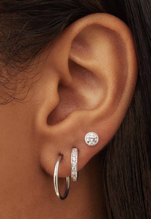 Boucles d'oreilles en argent, un anneau et un clou, exposées sur l'oreille ; le clou présente une pierre précieuse claire, tandis que l'anneau a une rangée horizontale de petites pierres.