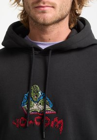 Zwarte hoodie met een kleurrijke geborduurde afbeelding van een groen wezen dat een boek leest. Kouwtjes aan de voorkant; zachte stoftextuur.