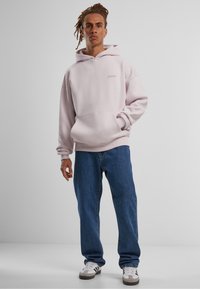 Haut à capuche rose clair avec une poche frontale et un logo sur la poitrine, associé à un jean bleu ample et des baskets blanches avec des accents gris.