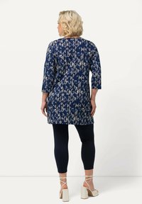 Granatowa tunika z geometrycznym kremowym wzorem, z rękawami o długości trzech ćwiartek, połączona z czarnymi legginsami i beżowymi sandałami na słupku.