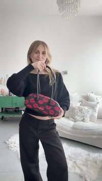 Mujer con atuendo negro sosteniendo un bolso negro con estampados de labios rojos, de pie en una habitación luminosa con un sofá blanco y un armario verde.