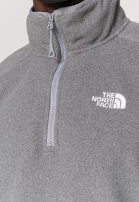 Suéter de felpa gris con cuello alto y cremallera frontal. Presenta un logo bordado en blanco en el lado izquierdo del pecho. Textura suave.