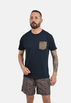 Uomo con barba e tatuaggio sulla manica che indossa una maglietta blu navy con tasca floreale e pantaloncini floreali abbinati su sfondo chiaro.