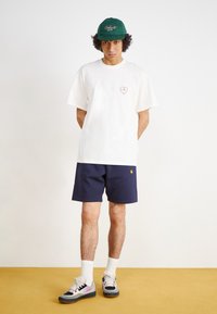 T-shirt en coton blanc avec un motif de cœur rouge, associé à un short bleu marine, des chaussettes blanches et des baskets multicolores, sur un fond jaune.
