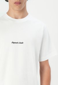Balta medvilnė marškinėliai su apvalia iškirpte ir trumpomis rankovėmis. Krūtinėje juodu siuvinėjimu užrašyta "Originals Studio". Lygus paviršius.