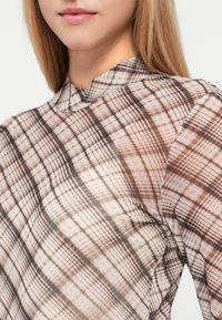Διαφανής μπλούζα με ψηλό γιακά, που διαθέτει καφέ και μπεζ καρό σχέδιο. Το ύφασμα έχει λεία υφή και ελαφριά εμφάνιση.