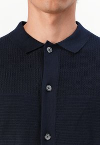 Marineblå strikket polo skjorte med teksturert øverste del, klassisk krage og tre knappelukking med mørke knapper.