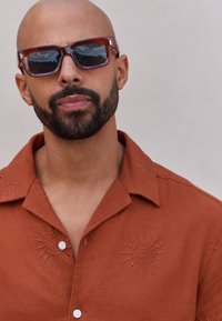 Brown linen shirt with sun embroidery on chest; white buttons; oversized rectangular sunglasses with a marbled brown frame and reflective lenses.