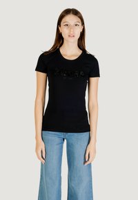 T-shirt nera in cotone con maniche corte, caratterizzata da un logo "GUESS" testurizzato in strass, abbinata a jeans a gamba larga di colore azzurro chiaro.