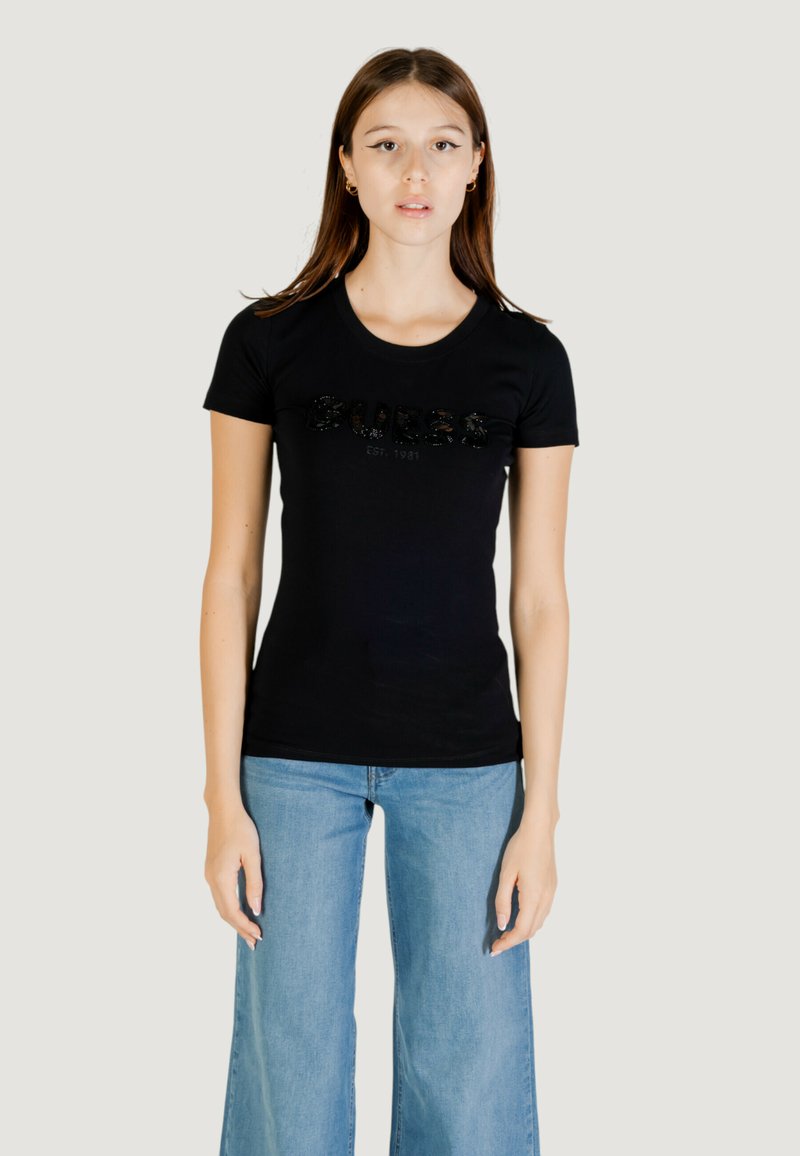 T-shirt nera in cotone con maniche corte, caratterizzata da un logo "GUESS" testurizzato in strass, abbinata a jeans a gamba larga di colore azzurro chiaro.