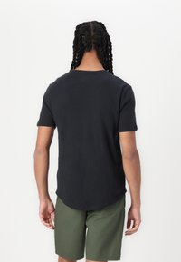 Schwarzes kurzärmliges T-Shirt aus leichtem Stoff mit abgerundetem Saum, kombiniert mit grünen Shorts, von hinten dargestellt.