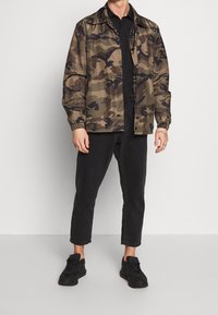 Giacca mimetica in tonalità oliva e marrone con polsini elastici e chiusura a bottoni a pressione, abbinata a jeans neri a gamba affusolata e sneakers nere.