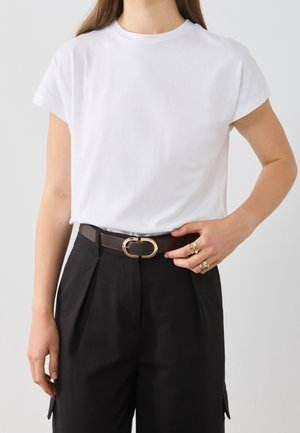Frau trägt ein schlichtes weißes T-Shirt mit kurzen Ärmeln, das in schwarze Hose mit hoher Taille gesteckt ist, mit einem dunkelbraunen Gürtel und goldener Schnalle.