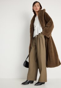 Casaco de pele falsa castanha, blusa branca com cordão, calças largas em verde-oliva e botins de salto pretos. Os acessórios incluem uma pequena mala preta.