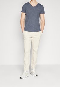 Modrý v-neck tričko s jemnou texturou, krémové kalhoty a světlé tenisky s síťovinovými akcenty a sportovním designem.