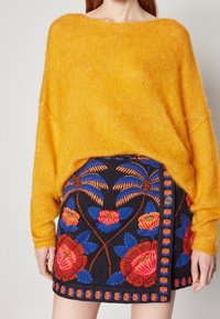 Gul, löst sittande tröja av mjukt material, kombinerad med en marinblå kjol med kraftig blomsterbroderi i röd, rosa och blå.