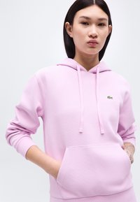 Νεαρό άτομο με ίσια μαύρα μαλλιά που φοράει μια ανοιχτόμωβ φούτερ κουκούλα με λογότυπο πράσινου κροκοδείλου και τατουάζ στο αντιβράχιο.