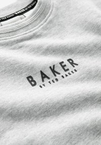 Siv gray sweatshirt z ribanim ovratnikom in dvignjenimi črnimi črkami, ki tvorijo napis "BAKER" s podnapisom "by TED BAKER". Mehka tekstura.