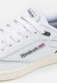 Bílé kožené tenisky Reebok s zelenými akcenty, s texturovaným povrchem, plochými tkaničkami a gumovou podrážkou s klasickým logem.