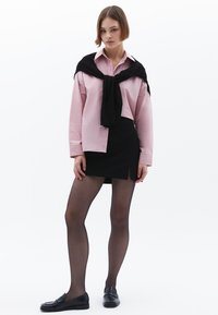 Lichtroze lange mouwen shirt met een witte kraag, gecombineerd met een zwarte mini-jurk en doorschijnende zwarte panty’s. Zwarte loafers maken de look compleet.
