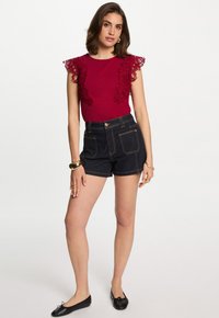 Top borgogna con dettagli in pizzo sulle spalle, abbinato a pantaloni corti in denim scuro con bottone frontale e tasche laterali. Indossati ballerine nere e accessori.