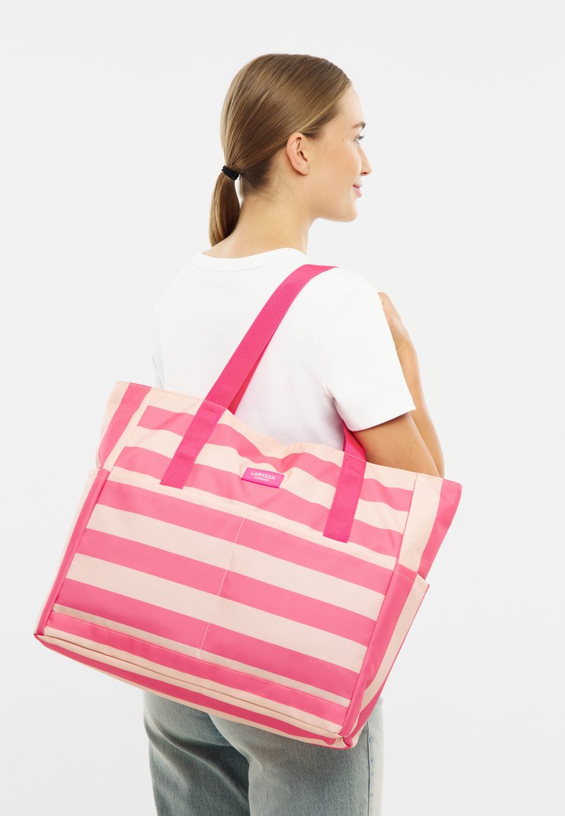 Kvinna med ljusbrunt hår i hästsvans bär en stor rosa och ljusrosa randig tote-väska över axeln, klädd i en vit skjorta och jeans.