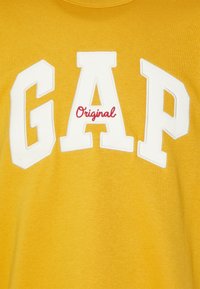 Camiseta de algodón amarilla con letras blancas en negrita "GAP" y un texto más pequeño en rojo que dice "Original" debajo. Tela suave y texturizada.