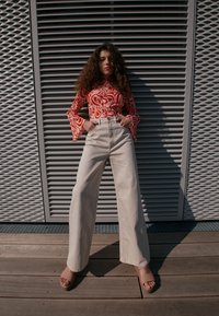 Jeune femme aux cheveux bouclés portant un haut rouge à motifs et un pantalon large beige taille haute, debout contre un mur métallique texturé.
