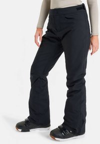 Mulher vestindo calças de esqui pretas e botas de neve pretas com solas brancas contra um fundo branco simples.