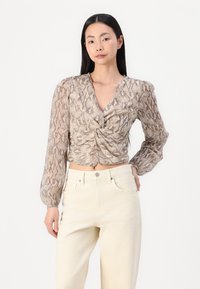 Blusa cropped con stampa serpente marrone e beige, scollo a V, dettaglio ancorato sul davanti e maniche lunghe a sbuffo, abbinata a jeans chiari.