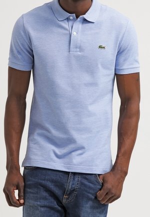 Mann, der ein hellblaues kurzärmeliges Polohemd mit kleinem grünem Krokodil-Logo und blaue Jeans trägt, steht mit einer Hand in der Tasche.