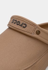 Gros plan sur un sabot Crocs couleur camel présentant une tige en tissu texturé et une bride ajustable avec le logo Crocs brodé en noir.