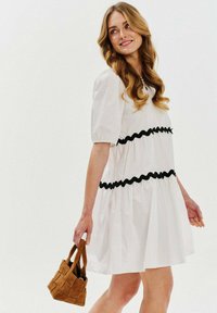 Biele krátke šaty s pufovanými rukávmi, s čiernym vlnitým lemovaním. Ďalším detailom je hnedá tkaná kabelka v ruke modelky.