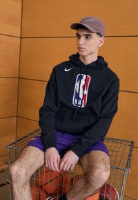 Hanorac negru cu logo-ul NBA, pantaloni scurți mov și o șapcă maro; așezat pe un cărucior metalic cu mingi de baschet dedesubt. Material neted, stil athletic.