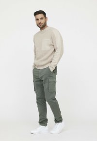 Beige gebreide trui met een zak, gecombineerd met olijfgroene cargo-broek met zijzakken. Draag met witte sportschoenen.