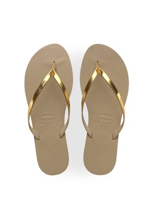 Tongs beiges texturées avec des lanières dorées brillantes en forme de V affichant le nom de la marque sur un fond blanc.