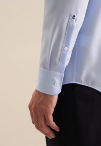 Lichtblauw overhemd met lange mouwen met twee witte knopen en een geborduurd bloemembleem dicht bij de manchetten. Glad stof met een lichte glans.