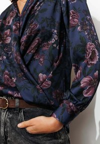 Blouse florale bleu nuit avec des fleurs roses et violettes, manches longues avec des poignets froncés, associée à un jean gris taille haute et une ceinture marron.