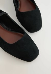 Ballerines en daim noir avec un bout carré et une fine lanière. La doublure intérieure est marron et la semelle extérieure est lisse. Design simple et élégant.