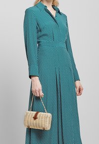Teal polka dot dress with a collar and long sleeves, featuring a fitted bodice and pleated skirt. Woven wicker handbag with a leather accent.