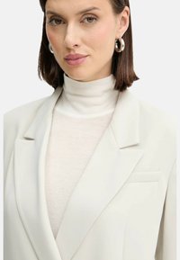 Blazer grigio chiaro con rever a punta, indossato sopra un dolcevita crema. Presenta una trama sottile e una tasca sul petto. Orecchini a cerchio in argento.