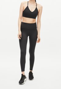 Kvinna iförd svart sport-bh, högmidjade svarta leggings och svarta löparskor stående framför en plain vit bakgrund.